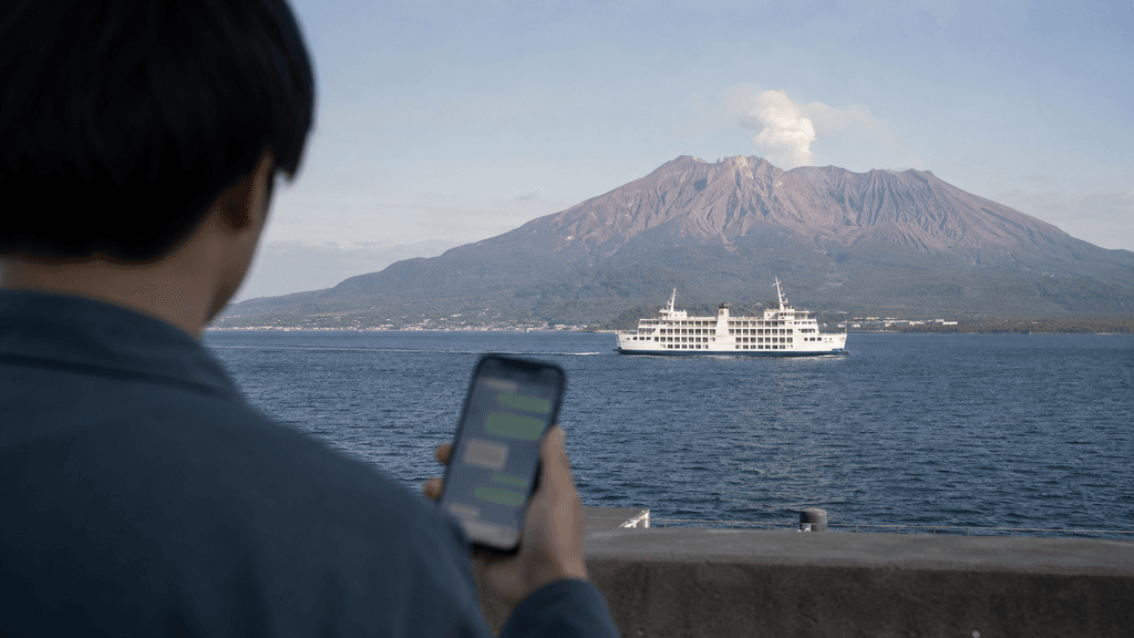 桜島を背景に錦江湾を渡るフェリーとスマートフォンの画面を見つめる男性のイメージ
